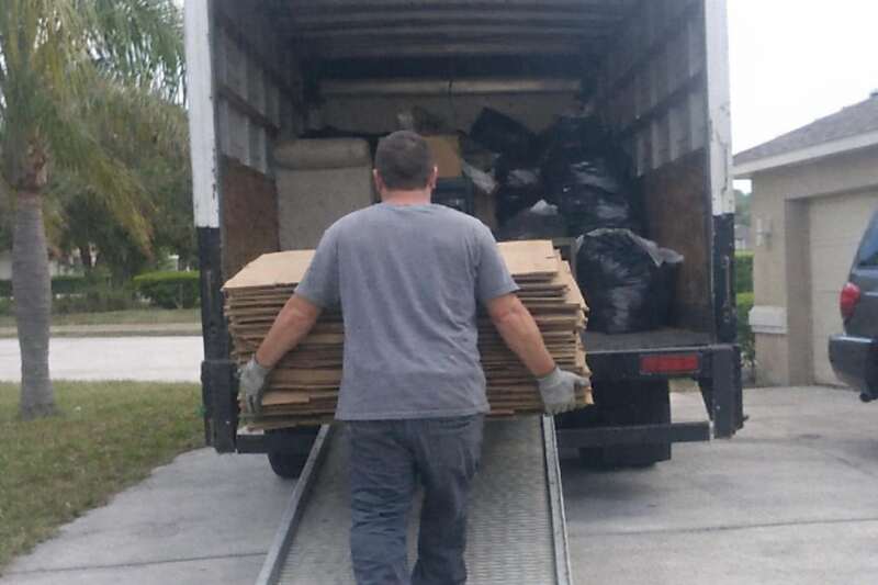 A worker seen from behind walking flattened cardboard up the ramp of a box truck