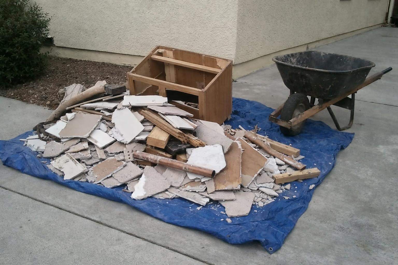 Renovation debris on a tarp at the edge of a driveway: broken tile, drywall offcuts, lumber and a torn-out vanity