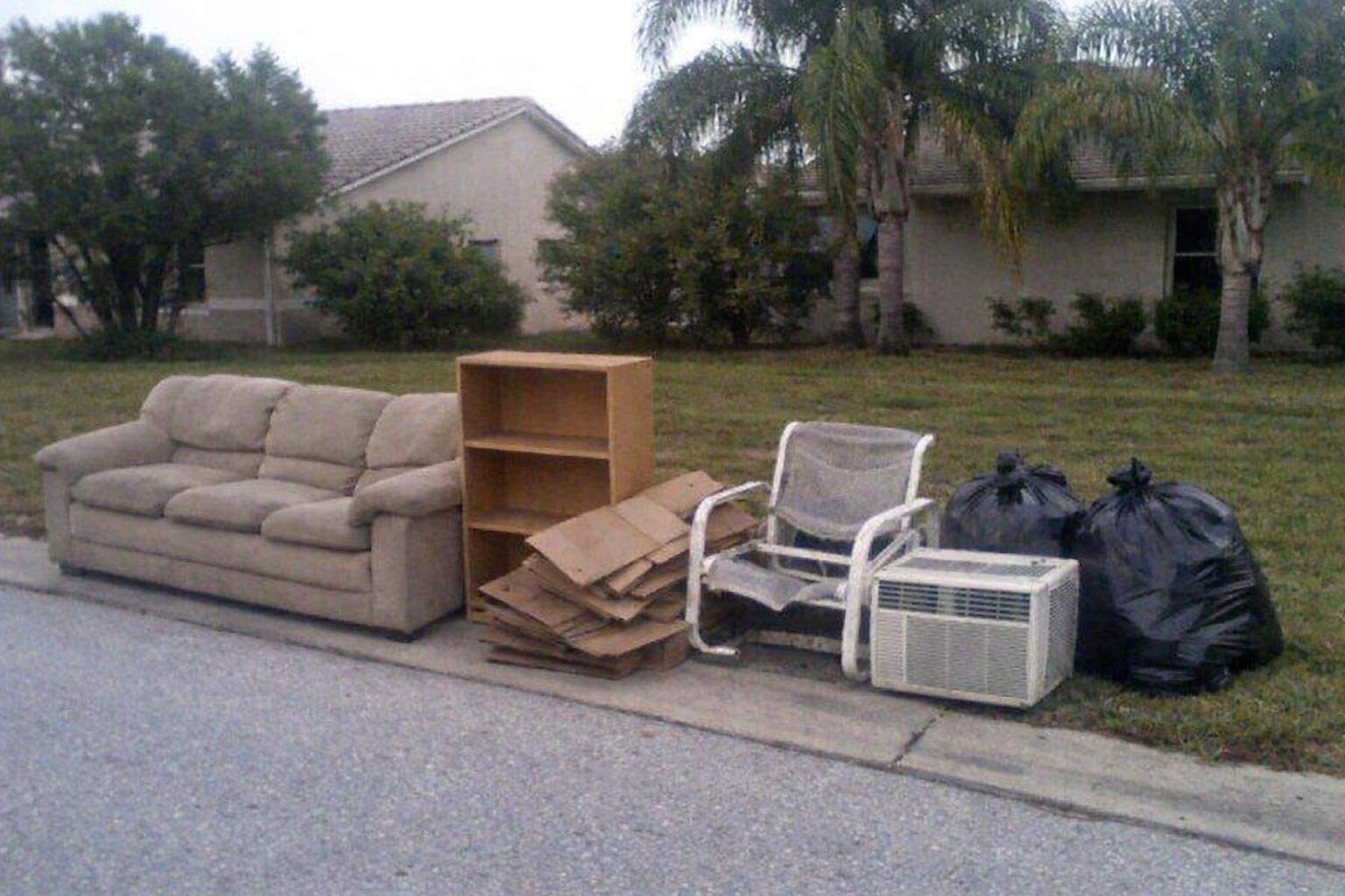A pile of mixed household junk at the edge of an Orlando driveway: a sofa, a bookcase, boxes and a window air conditioner
