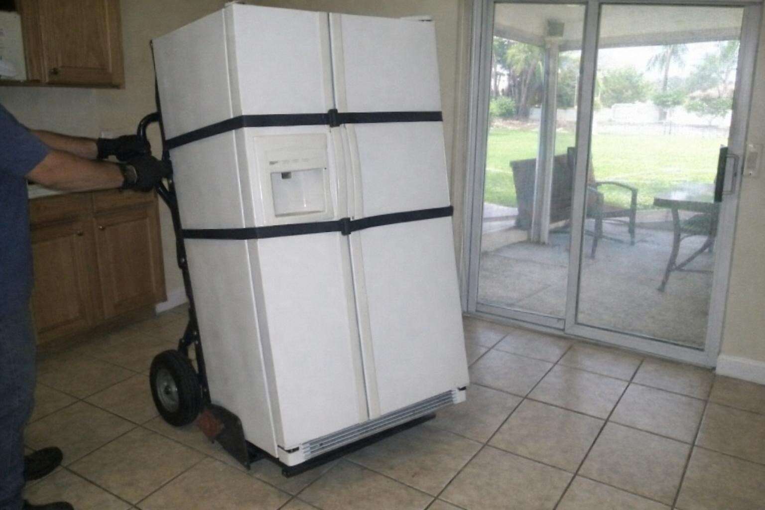 A refrigerator strapped to an appliance hand truck being wheeled across a tile kitchen floor