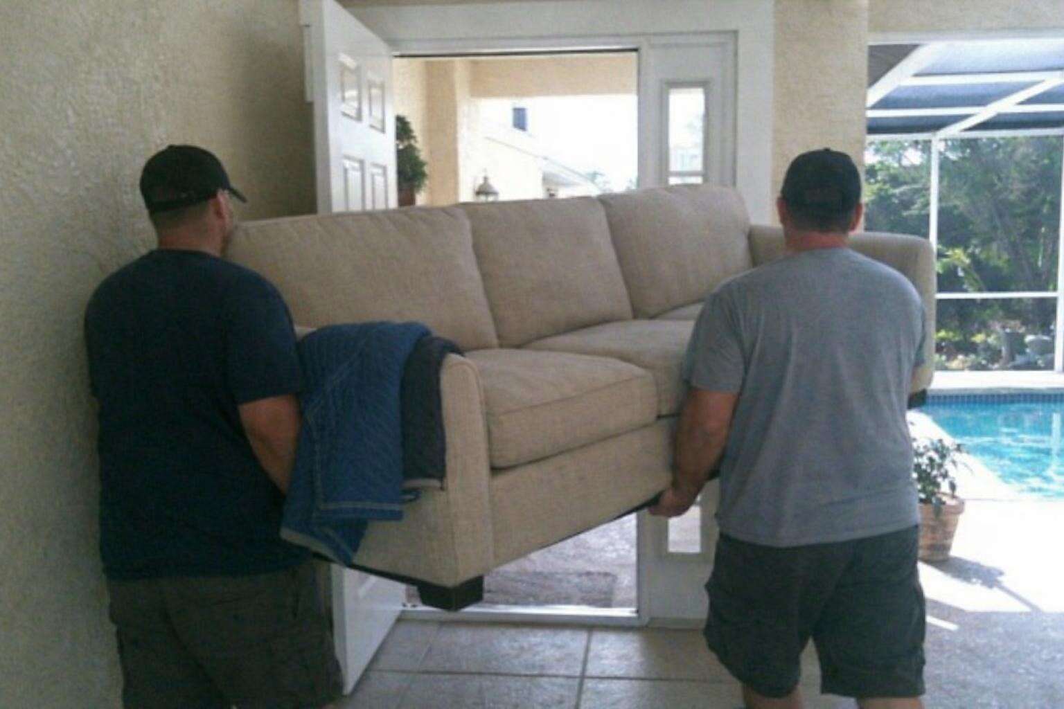 A three-seat sofa being carried sideways out through the front door of a stucco house